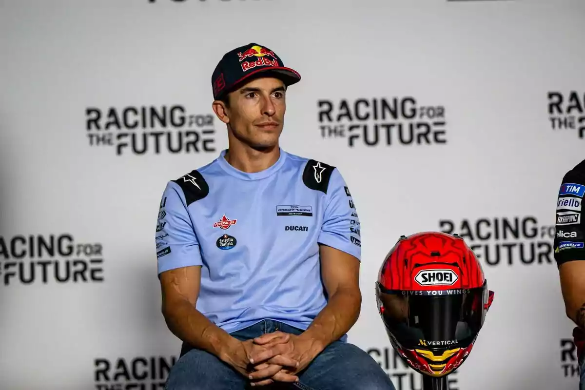 Un hombre con gorra y camiseta de equipo de motociclismo está sentado junto a un casco rojo en un evento con el fondo que dice Un hombre con gorra y camiseta de equipo de motociclismo está sentado junto a un casco rojo en un evento con el fondo que dice