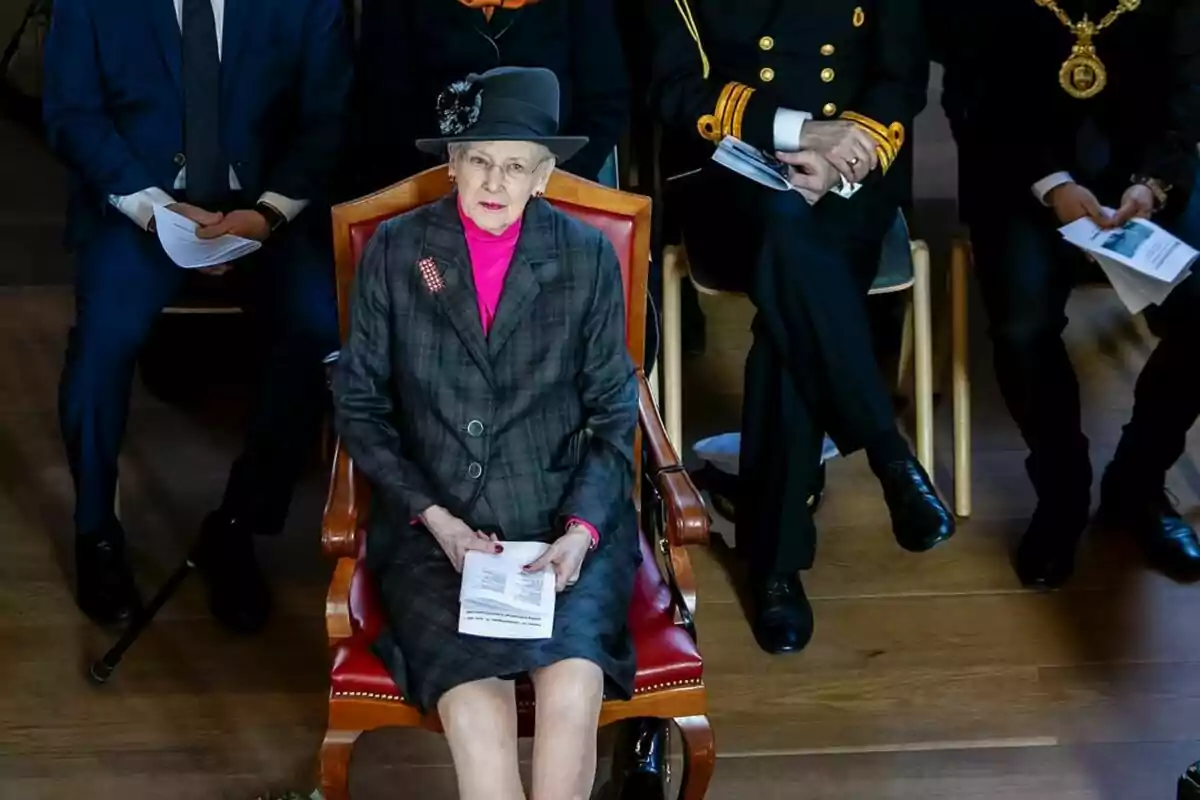 Una dona gran amb barret i abric fosc està asseguda en una cadira de fusta envoltada de persones vestides formalment. Una dona gran amb barret i abric fosc està asseguda en una cadira de fusta envoltada de persones vestides formalment.