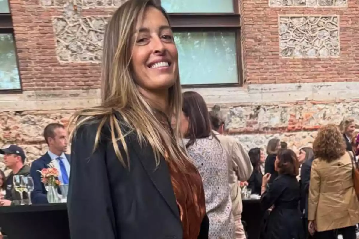 Una dona somrient amb cabell llarg i ros està dreta en un esdeveniment social a l'aire lliure, envoltada de persones conversant i gaudint de begudes, amb un edifici de maó al fons. Una dona somrient amb cabell llarg i ros està dreta en un esdeveniment social a l'aire lliure, envoltada de persones conversant i gaudint de begudes, amb un edifici de maó al fons.