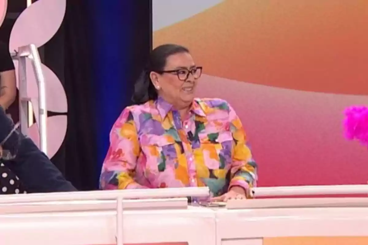 Una persona amb camisa acolorida i ulleres somriu mentre està asseguda en un plató de televisió amb fons acolorit. Una persona amb camisa acolorida i ulleres somriu mentre està asseguda en un plató de televisió amb fons acolorit.