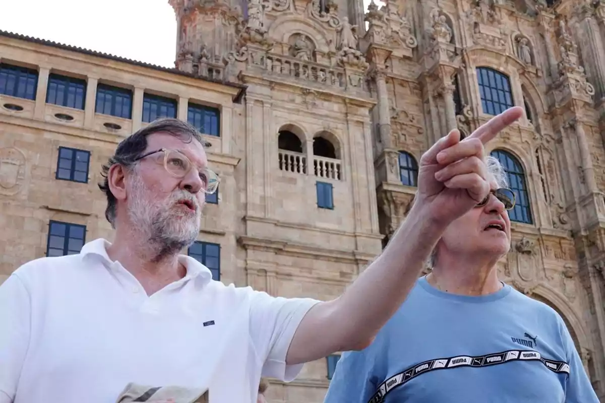 Dos homes grans observen i assenyalen alguna cosa a la distància davant d’un edifici històric de pedra amb finestres i detalls arquitectònics ornamentats. Dos homes grans observen i assenyalen alguna cosa a la distància davant d’un edifici històric de pedra amb finestres i detalls arquitectònics ornamentats.