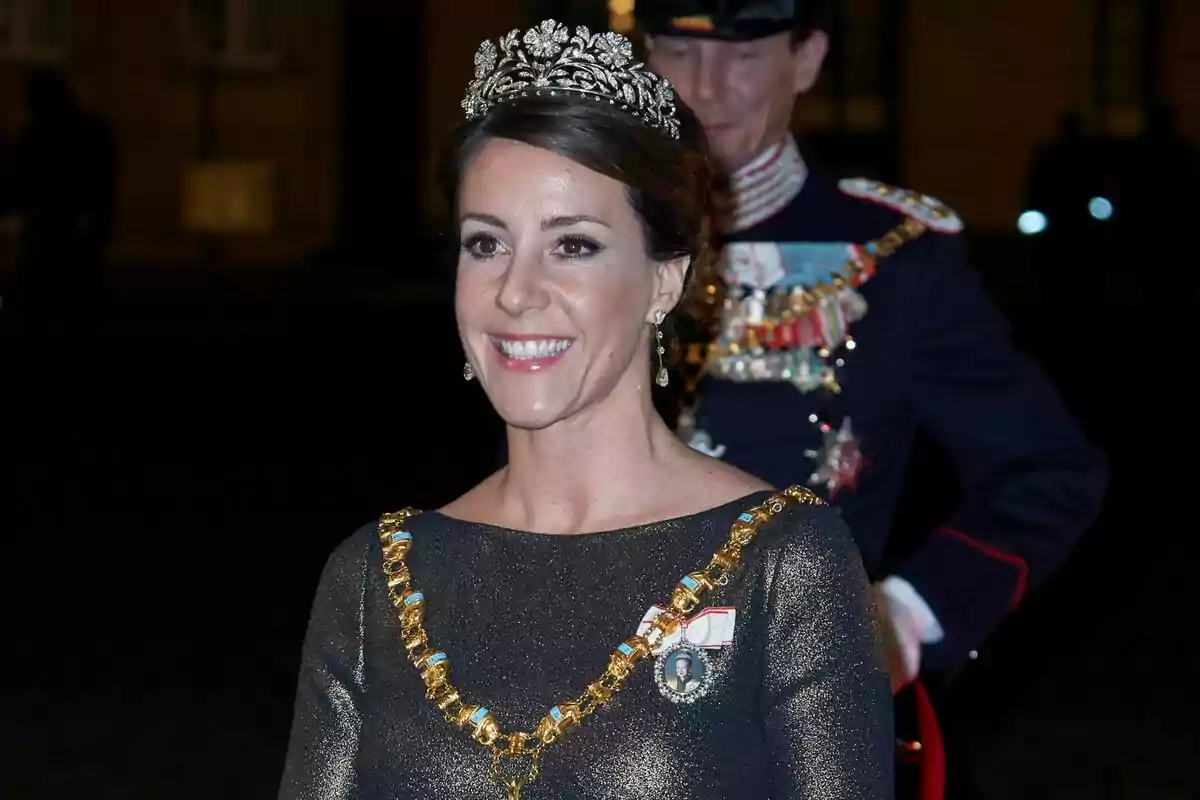 Una dona somrient porta una tiara i un collaret daurat en un esdeveniment formal. Una dona somrient porta una tiara i un collaret daurat en un esdeveniment formal.
