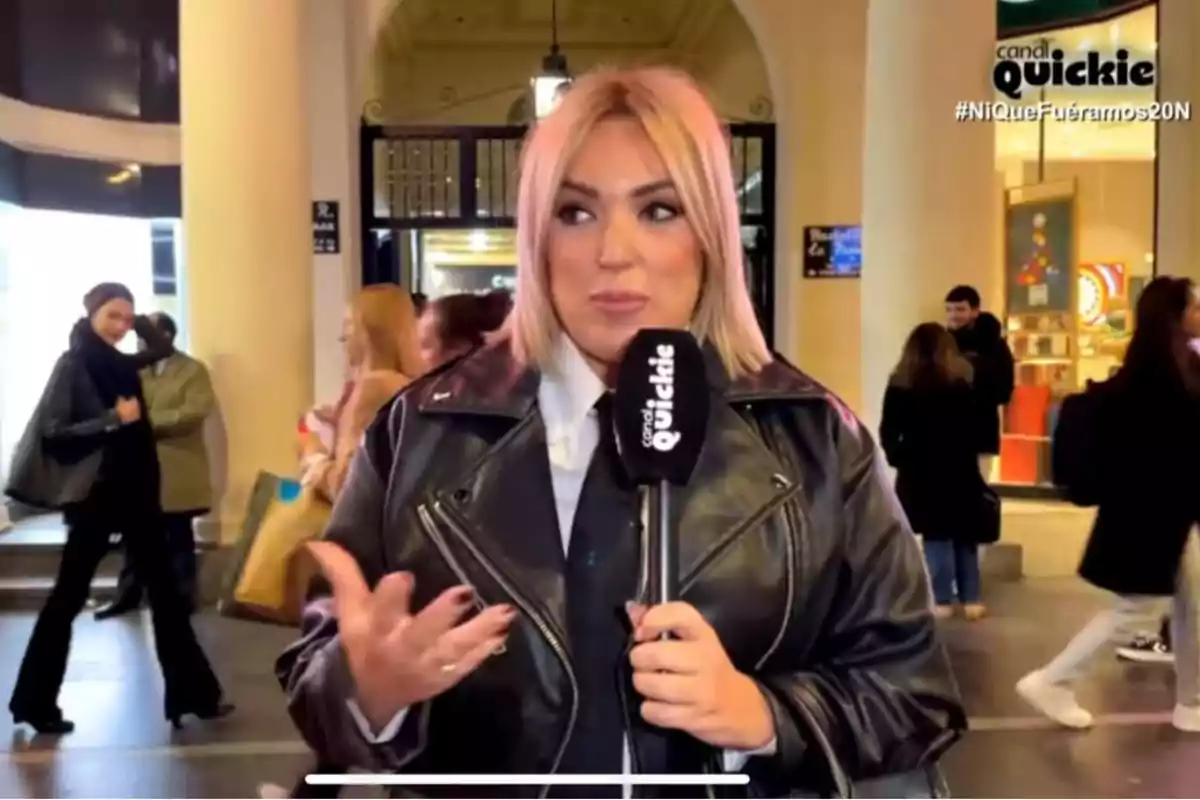 Una persona amb jaqueta de cuir sosté un micròfon en un lloc públic amb diverses persones caminant al fons.