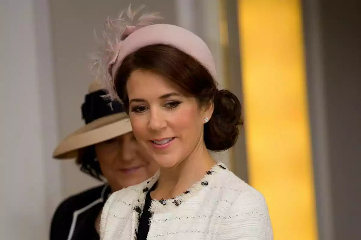 Una mujer con un elegante sombrero rosa y un peinado recogido sonríe mientras lleva un abrigo claro con detalles oscuros. Una mujer con un elegante sombrero rosa y un peinado recogido sonríe mientras lleva un abrigo claro con detalles oscuros.