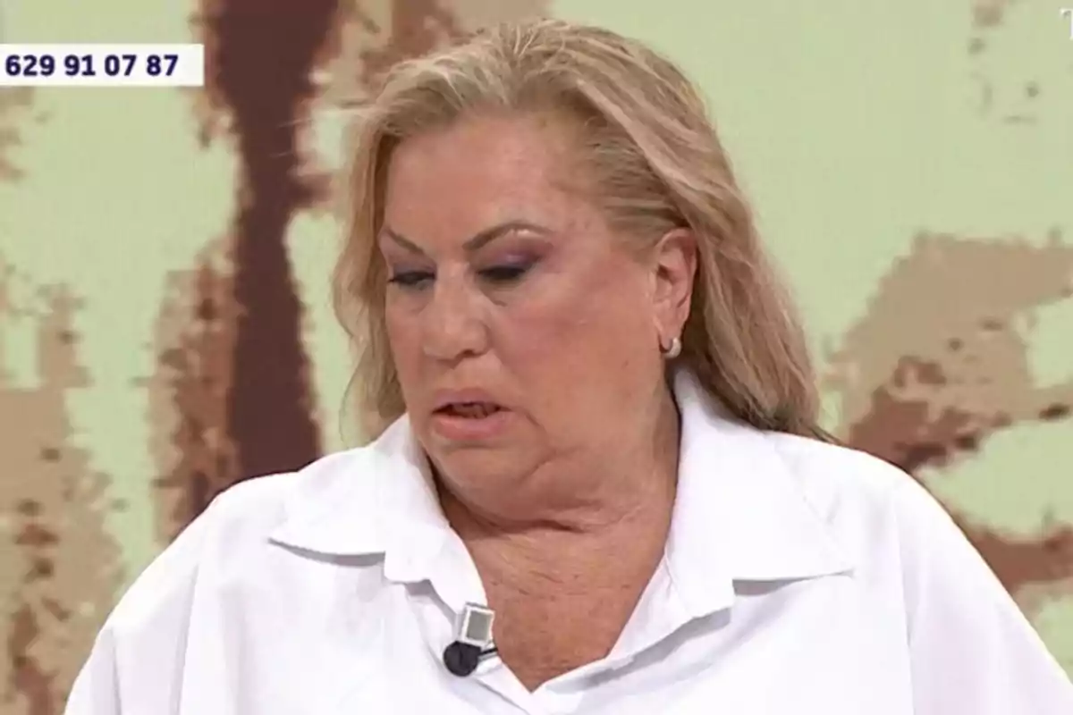 Una persona amb cabell ros i maquillatge pronunciat, vestint una camisa blanca i un micròfon de solapa, apareix en un entorn d'estudi de televisió amb un número de telèfon visible a la cantonada superior esquerra. Una persona amb cabell ros i maquillatge pronunciat, vestint una camisa blanca i un micròfon de solapa, apareix en un entorn d'estudi de televisió amb un número de telèfon visible a la cantonada superior esquerra.