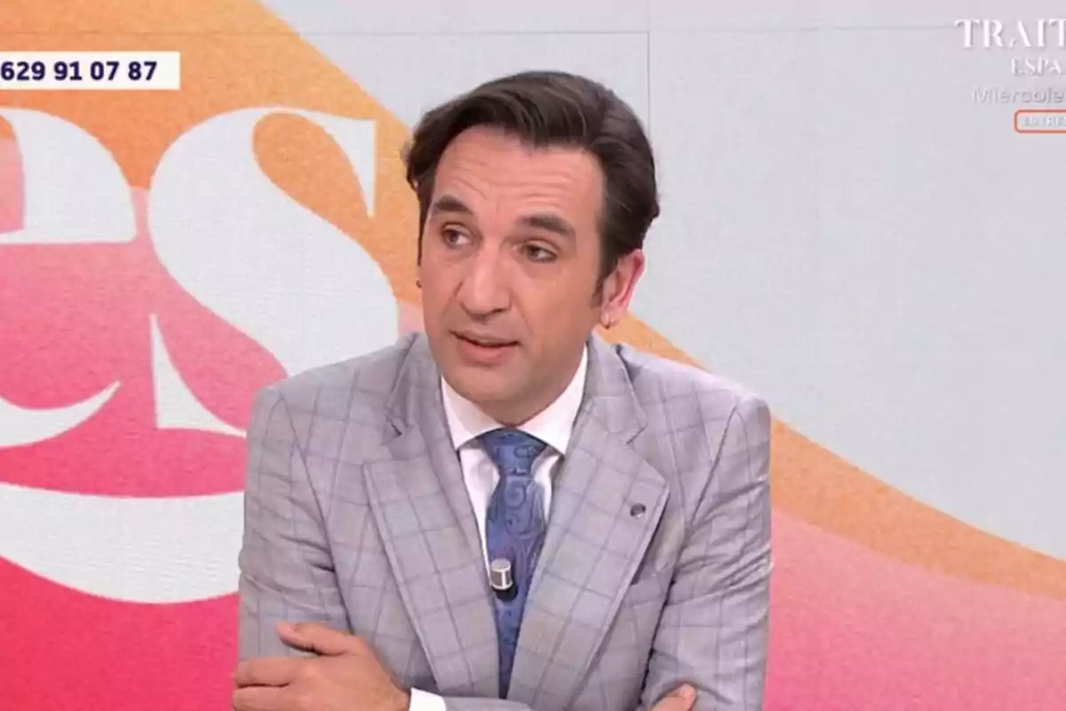 Un home amb vestit gris i corbata blava apareix en un programa de televisió amb un fons acolorit. Un home amb vestit gris i corbata blava apareix en un programa de televisió amb un fons acolorit.
