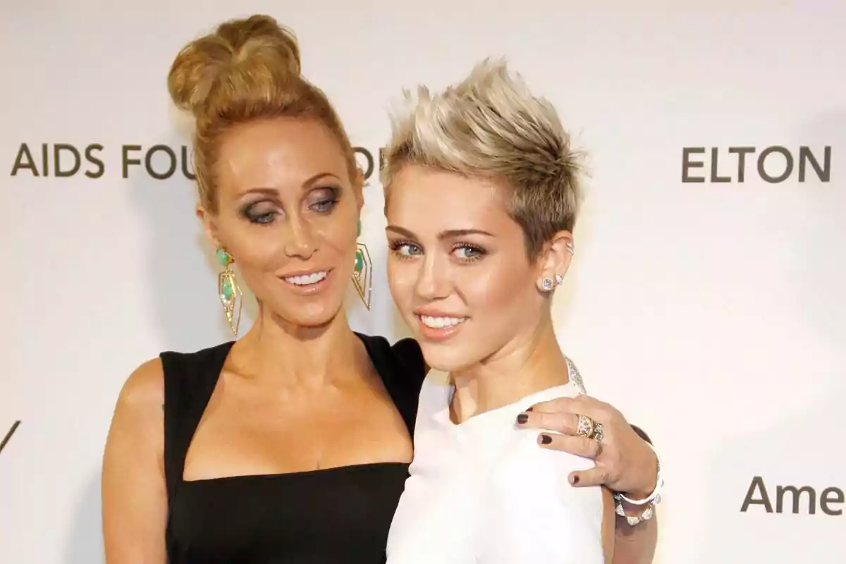 Two women pose together at an event with a white background and black letters; one is wearing a black dress and the other a white one. Both are smiling and look elegant.
