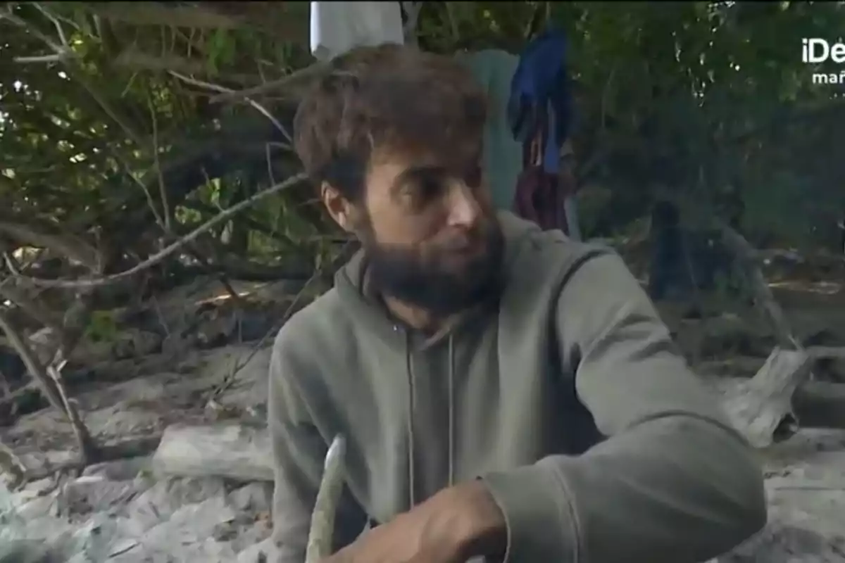 Un home amb barba i dessuadora està assegut en un entorn natural envoltat de branques i vegetació. Un home amb barba i dessuadora està assegut en un entorn natural envoltat de branques i vegetació.