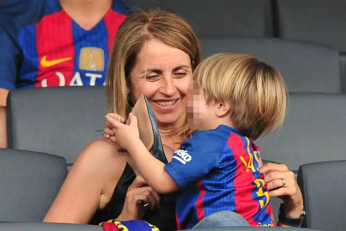 Montserrat Bernabeu sonríe mientras sostiene a Sasha Piqué que lleva una camiseta de fútbol azul y roja.