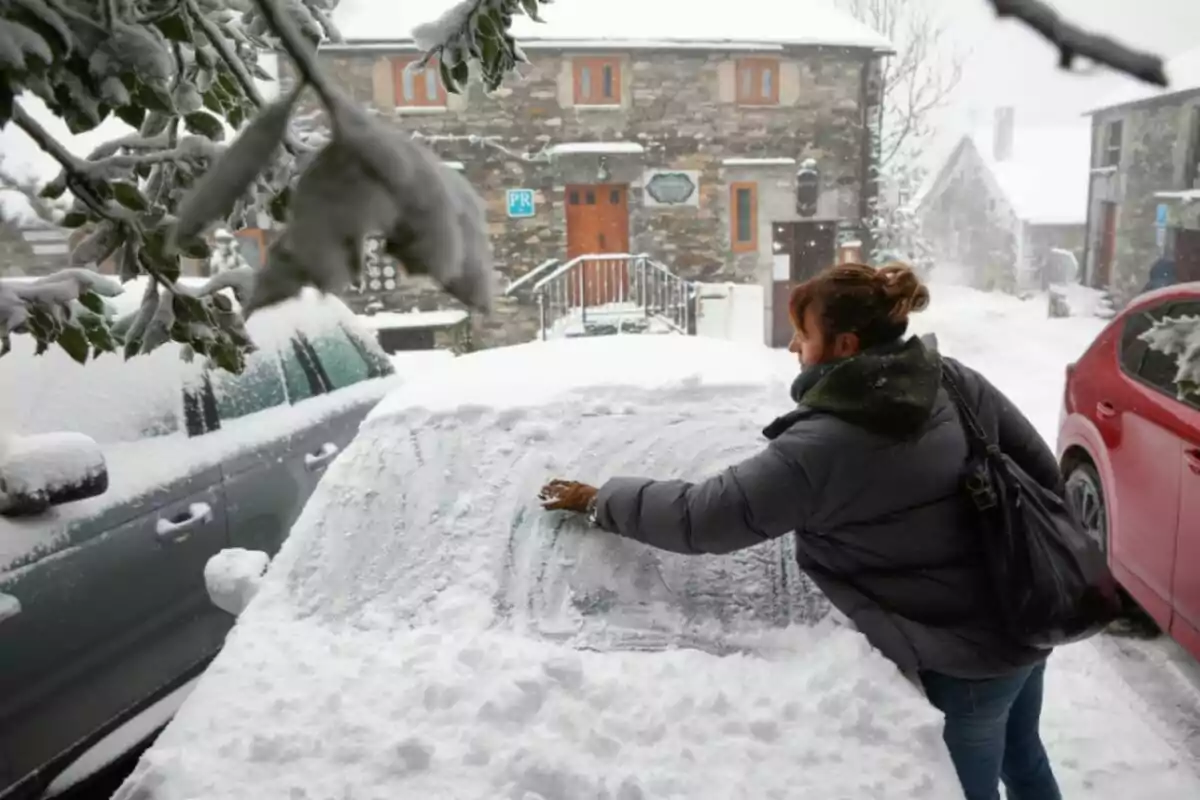 Una persona neteja la neu acumulada sobre un automòbil en un entorn nevat amb edificis de pedra al fons.