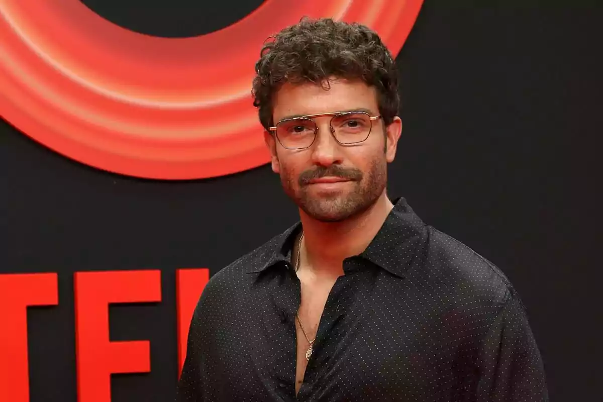 Home amb ulleres i camisa negra posant davant d’un fons amb el logotip de Netflix en tons vermells.