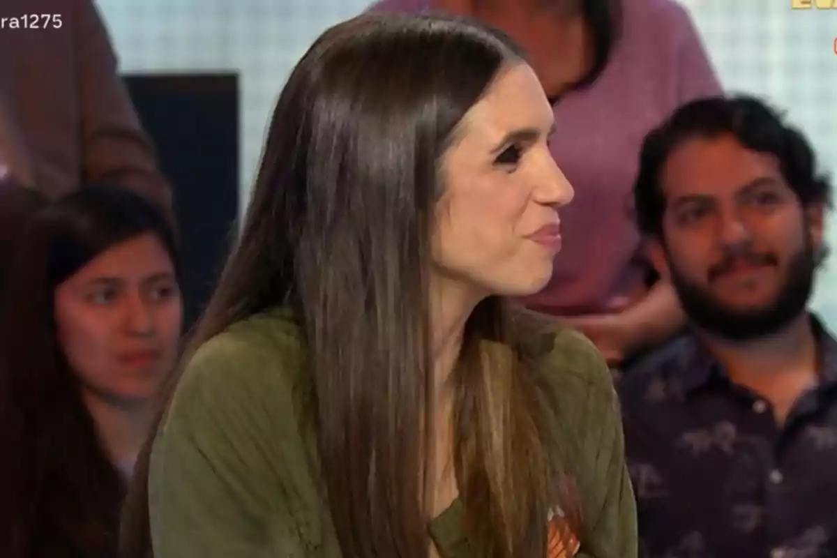 Una dona de cabell llarg i fosc està asseguda de perfil en un entorn d'estudi amb persones al fons. Una dona de cabell llarg i fosc està asseguda de perfil en un entorn d'estudi amb persones al fons.