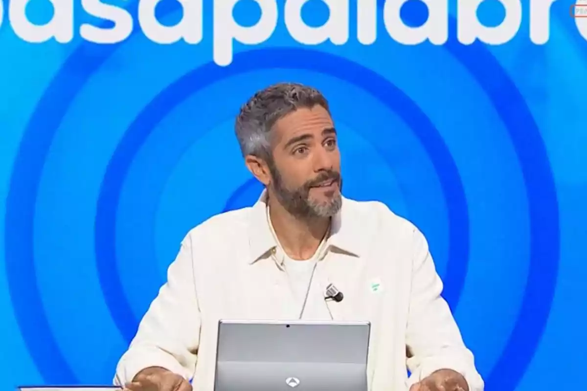 Home de cabells curts i barba assegut davant d’un ordinador en un plató de televisió amb fons blau i lletres blanques Home de cabells curts i barba assegut davant d’un ordinador en un plató de televisió amb fons blau i lletres blanques