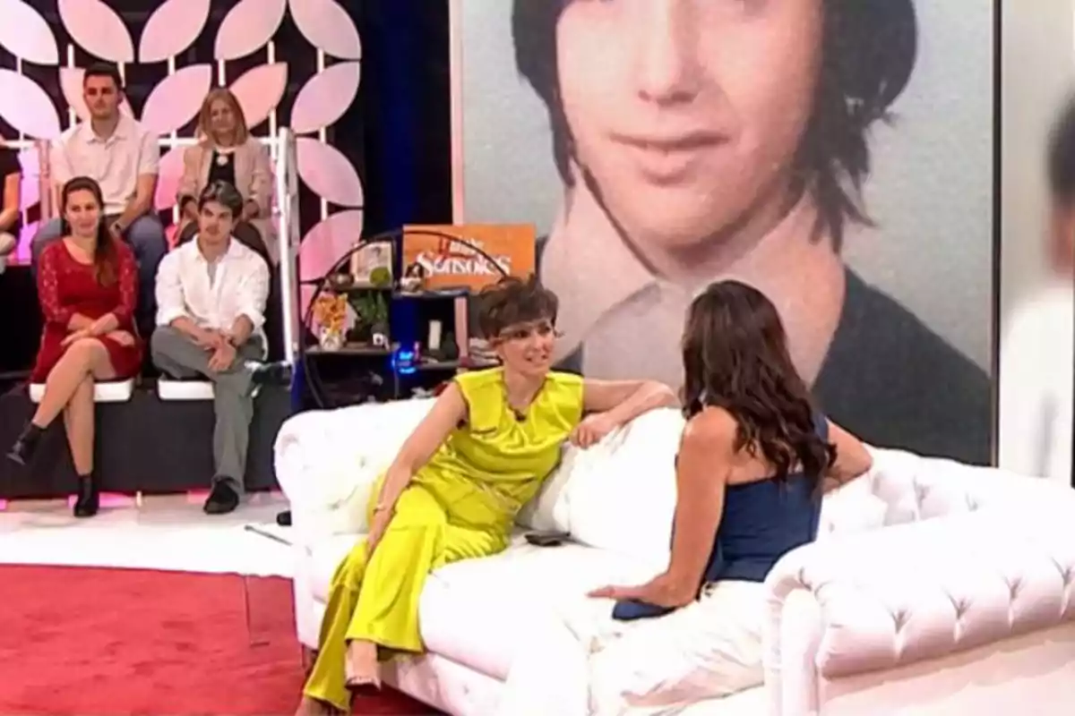 Un grup de persones és en un plató de televisió, dues dones conversen en un sofà blanc mentre altres persones estan assegudes al fons, i una gran imatge d'una persona jove es mostra a la pantalla darrere d'elles. Un grup de persones és en un plató de televisió, dues dones conversen en un sofà blanc mentre altres persones estan assegudes al fons, i una gran imatge d'una persona jove es mostra a la pantalla darrere d'elles.