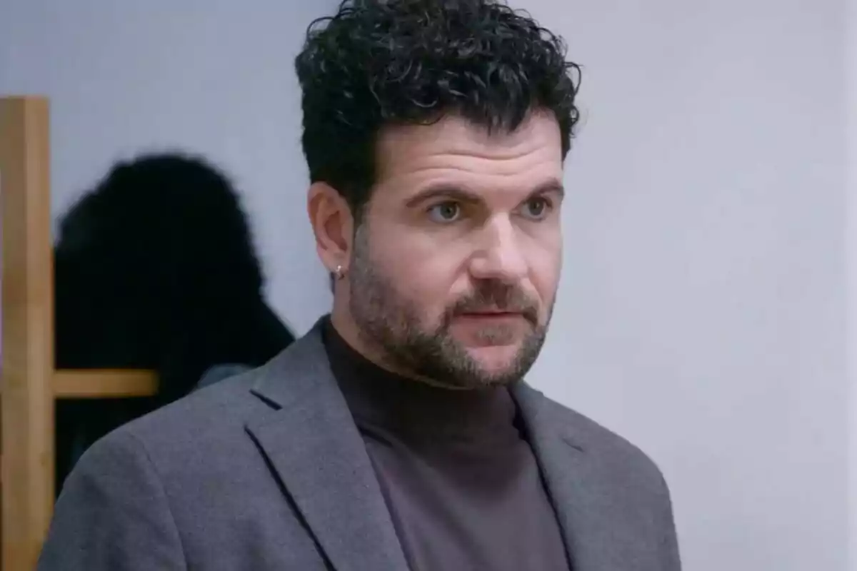 Un hombre con barba y cabello rizado lleva un pendiente y viste un suéter oscuro con un saco gris. Un hombre con barba y cabello rizado lleva un pendiente y viste un suéter oscuro con un saco gris.