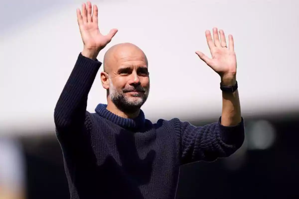 A bald man with a beard and a dark sweater raises both hands in greeting. A bald man with a beard and a dark sweater raises both hands in greeting.