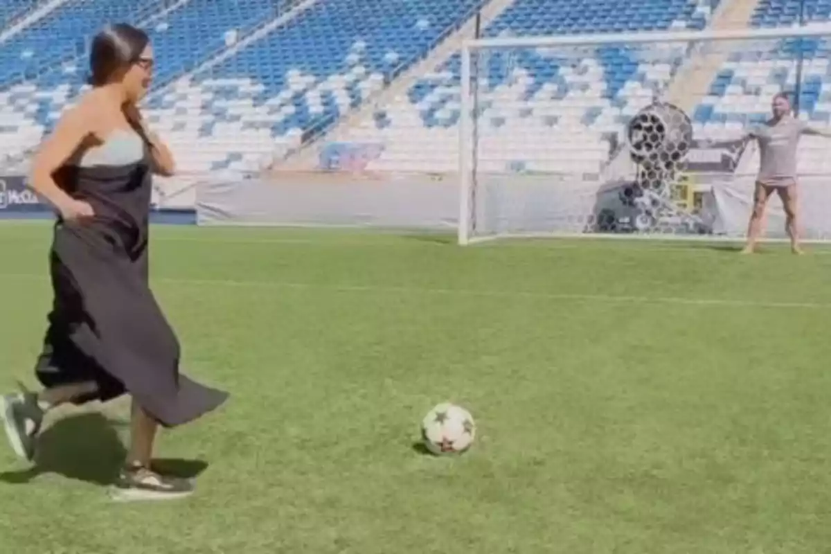 Una dona amb vestit negre i sabatilles es prepara per xutar una pilota de futbol en un estadi mentre una persona està a la porteria. Una dona amb vestit negre i sabatilles es prepara per xutar una pilota de futbol en un estadi mentre una persona està a la porteria.
