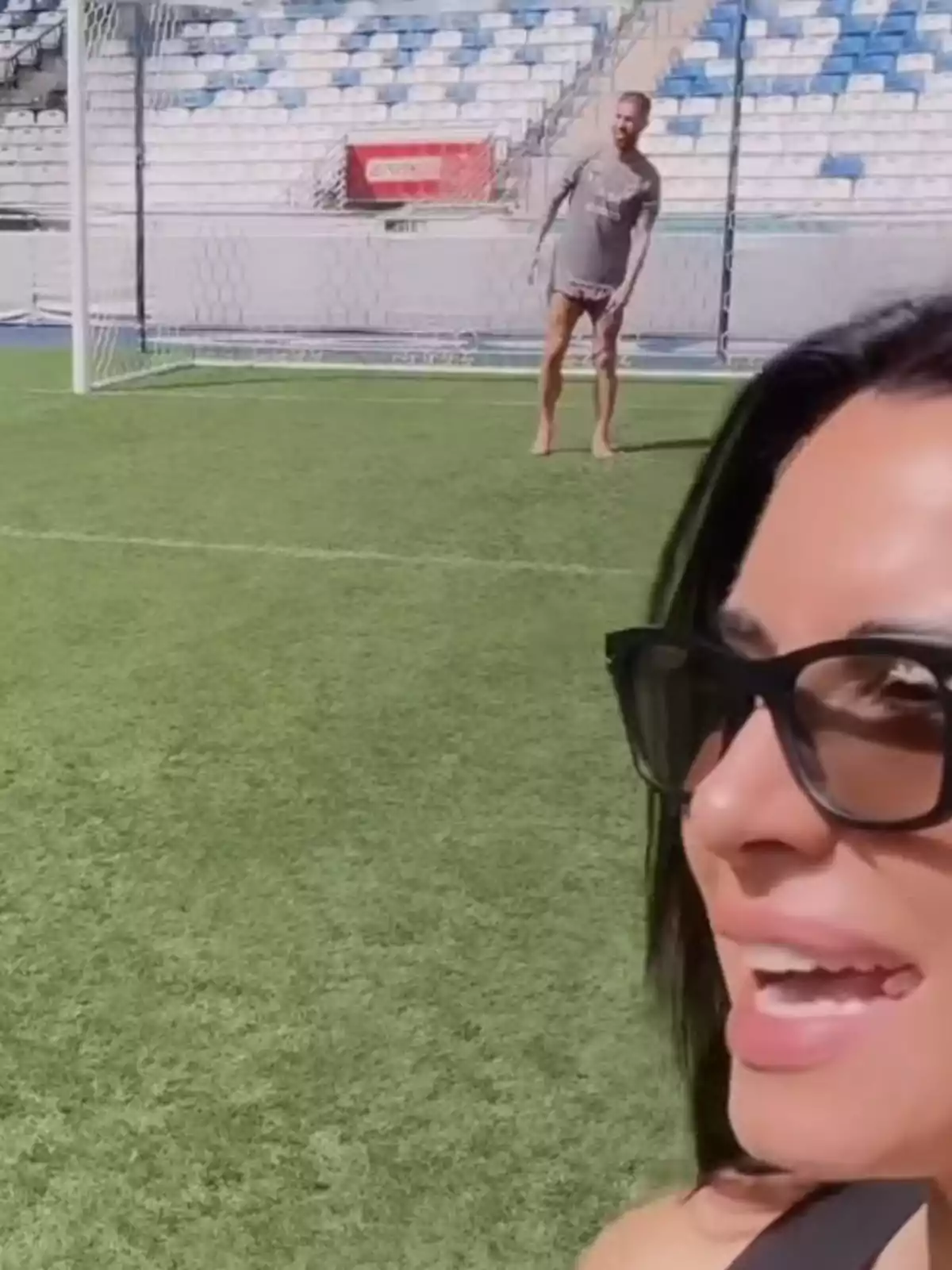 Una dona amb ulleres de sol somriu en primer pla mentre un home està descalç en una porteria de futbol al fons. Una dona amb ulleres de sol somriu en primer pla mentre un home està descalç en una porteria de futbol al fons.