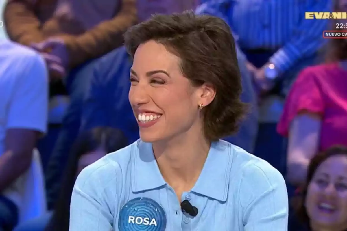 Dona somrient amb cabell curt i camisa blau clar participant en un programa de televisió amb públic de fons i una placa amb el nom Rosa al pit. Dona somrient amb cabell curt i camisa blau clar participant en un programa de televisió amb públic de fons i una placa amb el nom Rosa al pit.