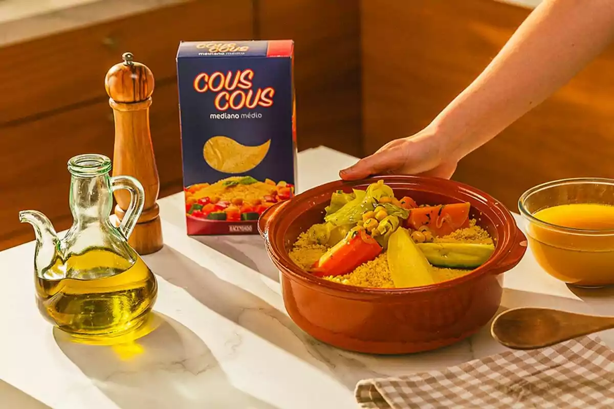 Un plat de cuscús amb verdures en un recipient de fang al costat d'una caixa de cuscús, una ampolla d'oli d'oliva, un molinet de pebre i un bol de brou sobre una taula. Un plat de cuscús amb verdures en un recipient de fang al costat d'una caixa de cuscús, una ampolla d'oli d'oliva, un molinet de pebre i un bol de brou sobre una taula.
