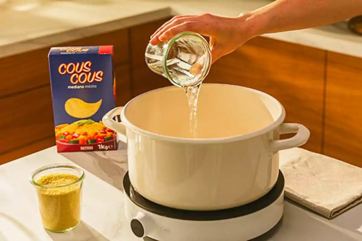 Una mà abocant aigua en una olla blanca sobre una estufa, amb una caixa de cuscús i un pot de cuscús a la taula. Una mà abocant aigua en una olla blanca sobre una estufa, amb una caixa de cuscús i un pot de cuscús a la taula.