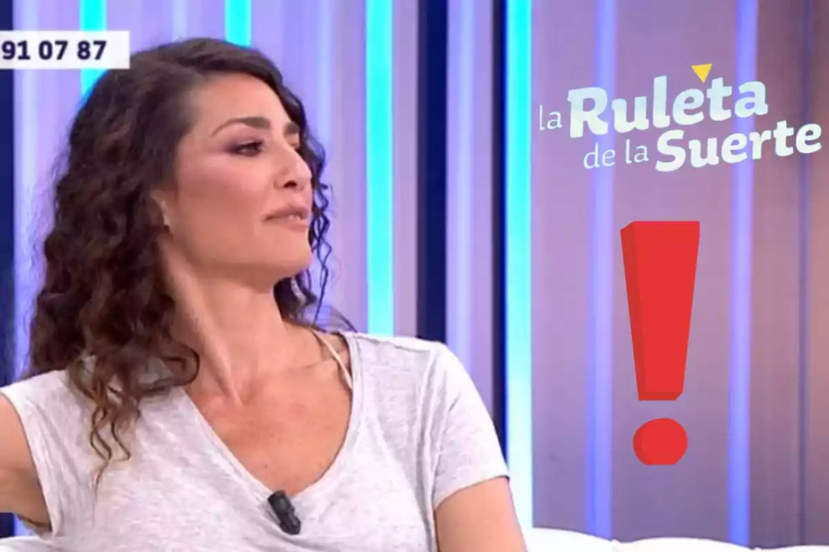 Dona de cabells arrissats i samarreta clara asseguda en un plató de televisió amb el logotip de La Ruleta de la Suerte i un signe d’exclamació vermell al seu costat