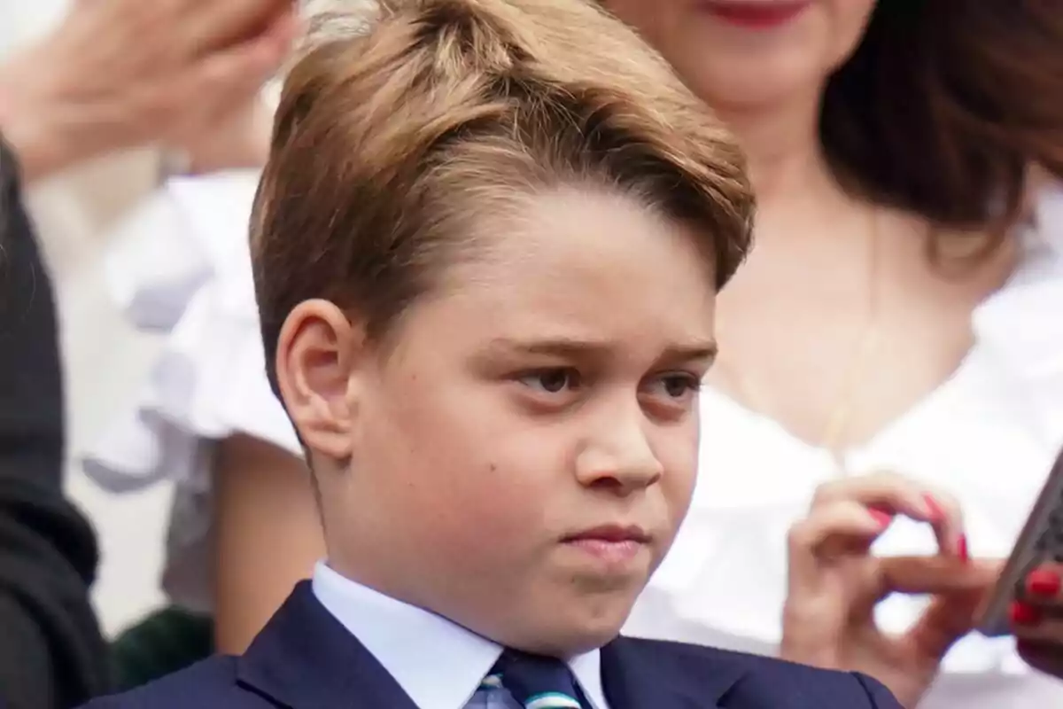 Un nen amb expressió seriosa, vestit amb vestit i corbata, envoltat de persones en un esdeveniment. Un nen amb expressió seriosa, vestit amb vestit i corbata, envoltat de persones en un esdeveniment.