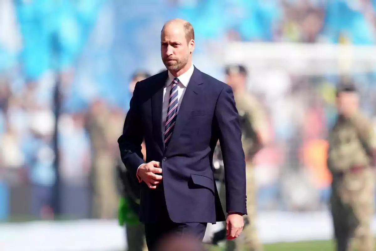 A man in a dark suit and tie walks at an outdoor event with uniformed people in the background. A man in a dark suit and tie walks at an outdoor event with uniformed people in the background.