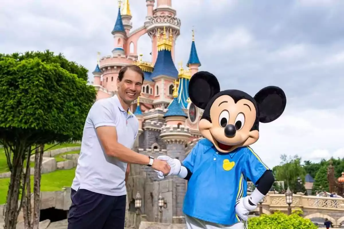 Un home somrient estreny la mà de Mickey Mouse davant d’un castell en un parc temàtic Un home somrient estreny la mà de Mickey Mouse davant d’un castell en un parc temàtic