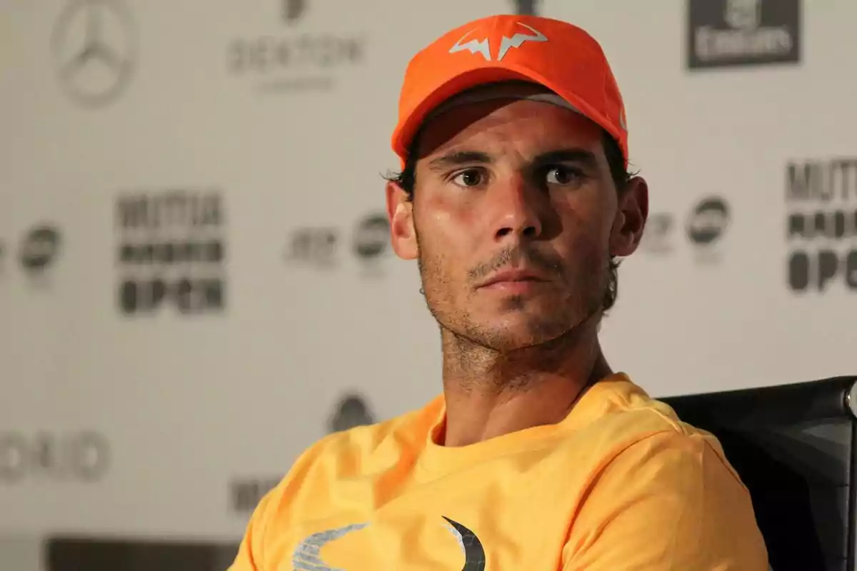 Hombre con gorra y camiseta naranja sentado en una conferencia de prensa de tenis