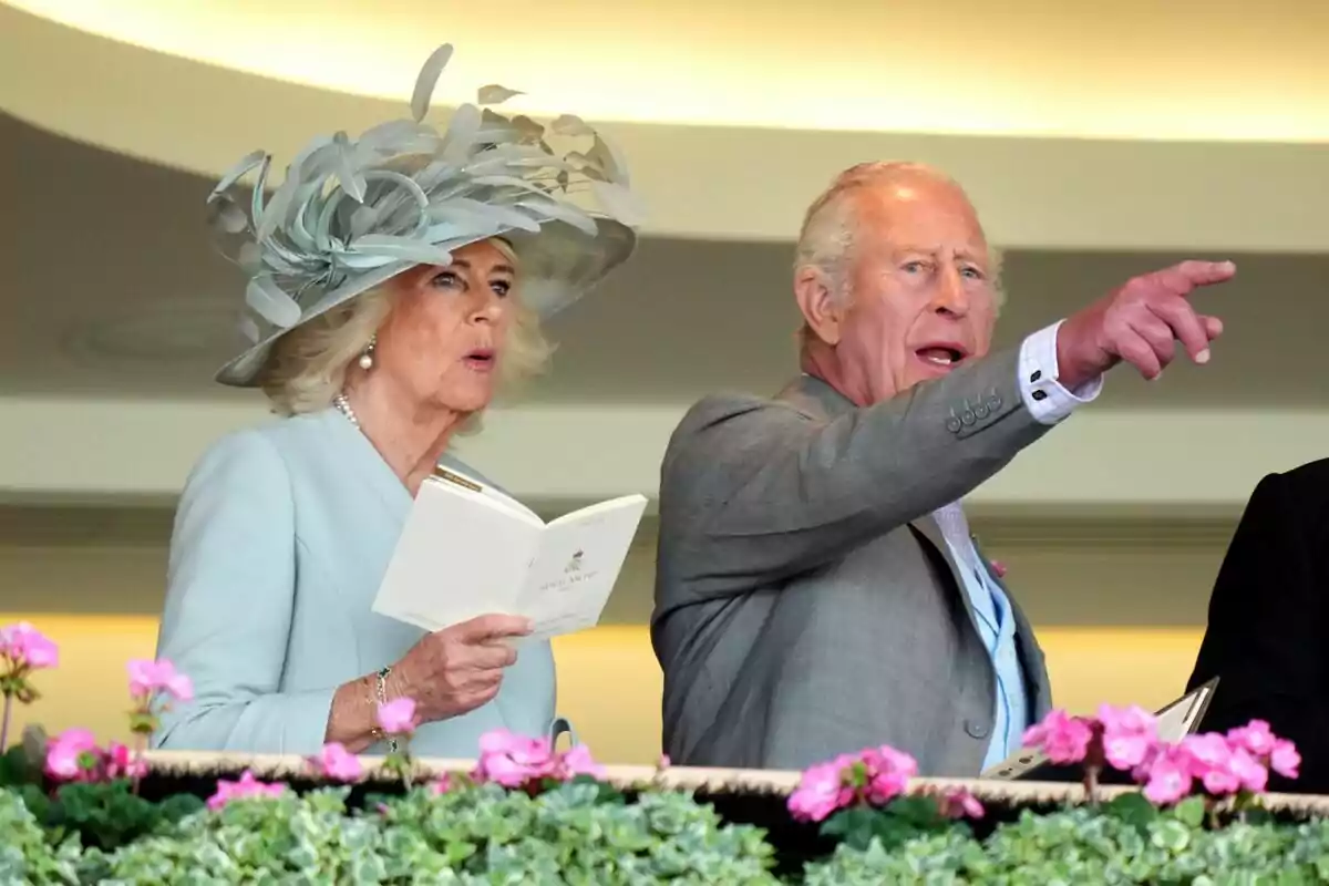 Una dona elegantment vestida amb un gran barret sosté un programa mentre un home al seu costat assenyala cap al davant envoltats de flors roses. Una dona elegantment vestida amb un gran barret sosté un programa mentre un home al seu costat assenyala cap al davant envoltats de flors roses.