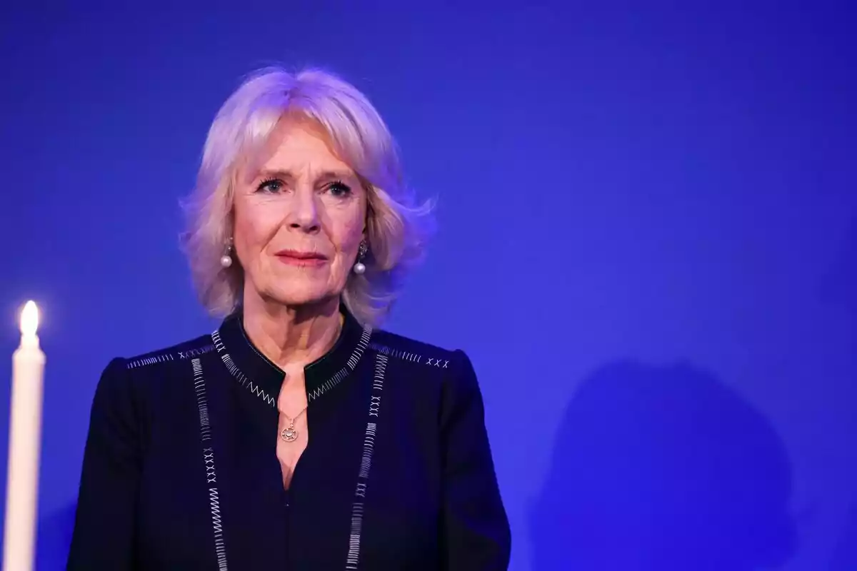 Blonde, short-haired woman with a serious expression dressed in black, standing in front of a blue background with a lit candle beside her Blonde, short-haired woman with a serious expression dressed in black, standing in front of a blue background with a lit candle beside her