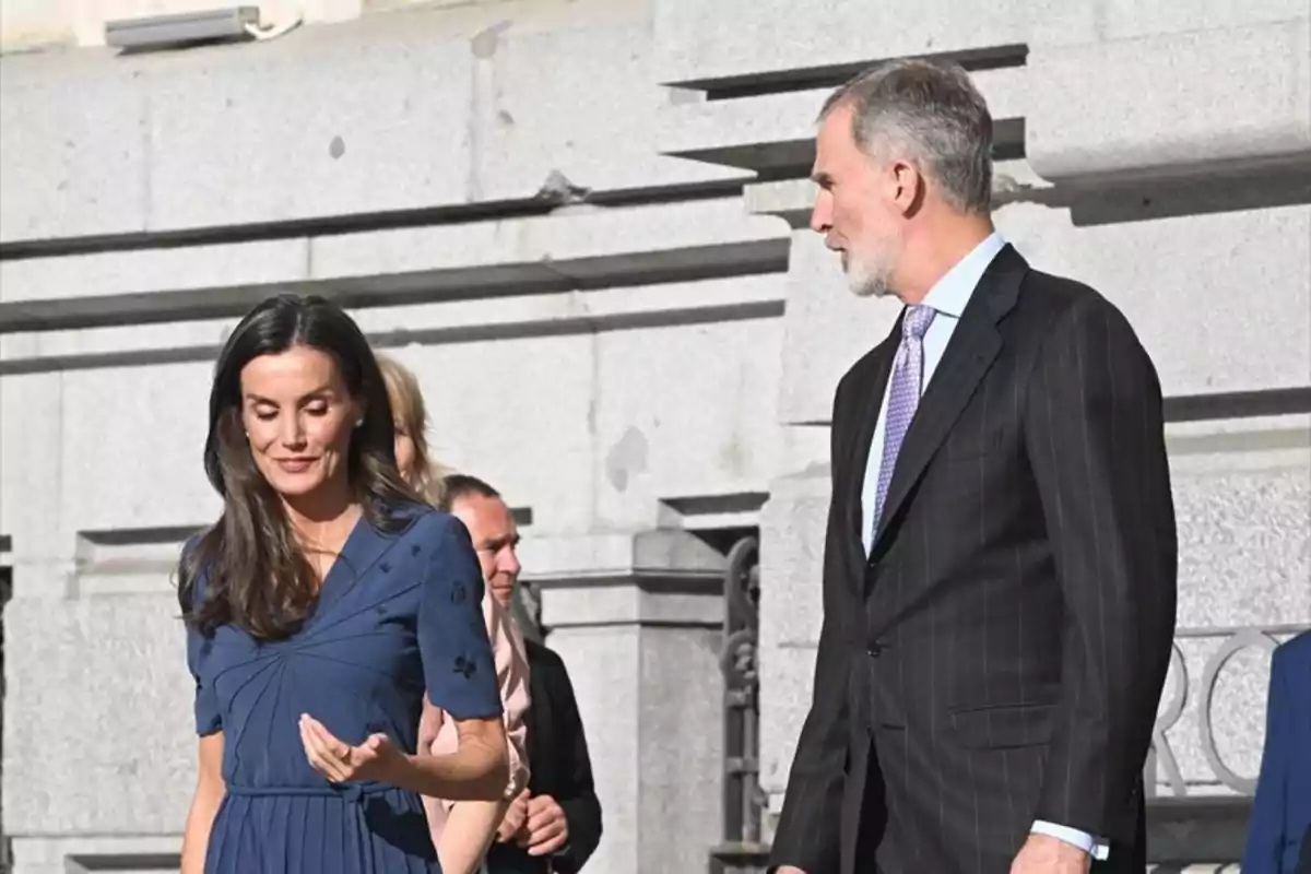 Una dona amb un vestit blau i un home amb vestit fosc caminen junts davant d'un edifici de pedra. Una dona amb un vestit blau i un home amb vestit fosc caminen junts davant d'un edifici de pedra.