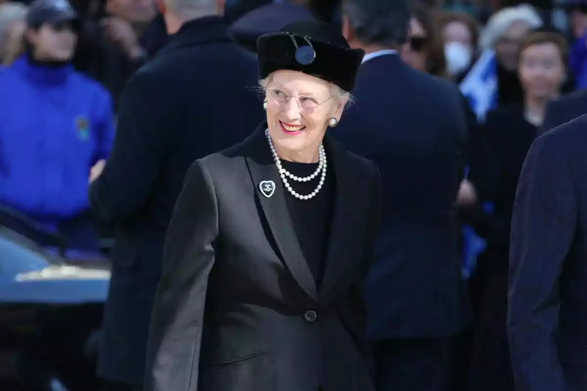 An older woman with glasses and a black hat smiles while wearing an elegant black outfit with a pearl necklace, surrounded by people at an outdoor event. An older woman with glasses and a black hat smiles while wearing an elegant black outfit with a pearl necklace, surrounded by people at an outdoor event.