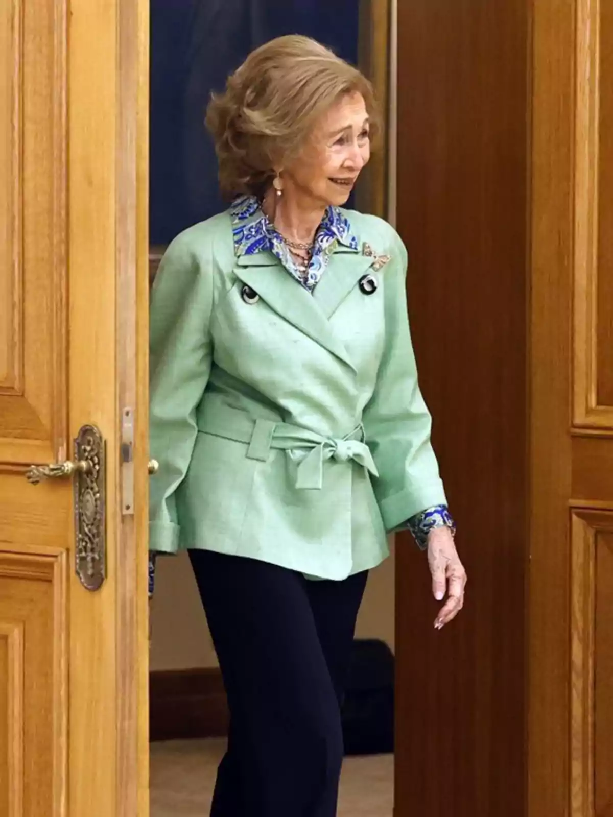 Dona gran amb jaqueta verd clar i pantalons foscos entrant per una porta de fusta Dona gran amb jaqueta verd clar i pantalons foscos entrant per una porta de fusta