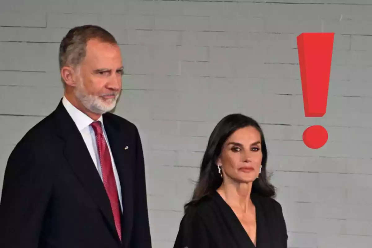 Un home i una dona elegantment vestits posen davant d’una paret grisa amb un gran signe d’exclamació vermell al seu costat.