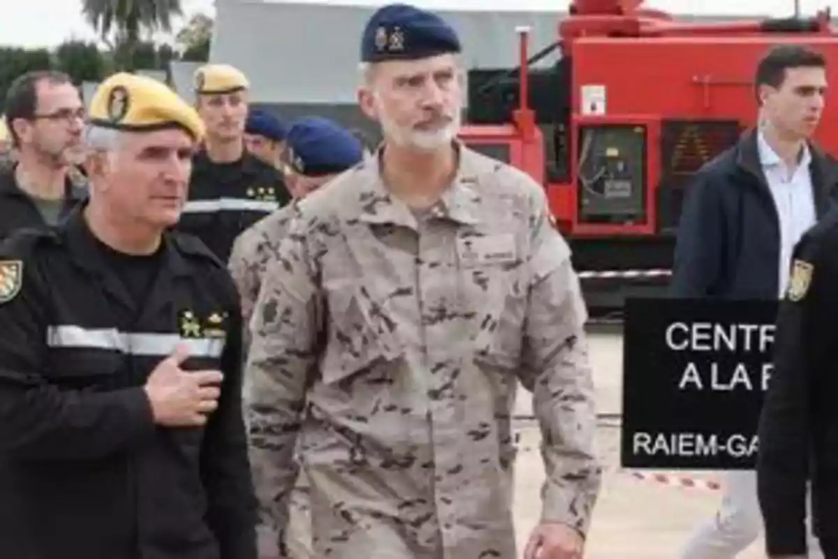 Un grup de persones amb uniformes militars i boines de diferents colors caminen a l'aire lliure a prop d'un edifici i un vehicle vermell. Un grup de persones amb uniformes militars i boines de diferents colors caminen a l'aire lliure a prop d'un edifici i un vehicle vermell.