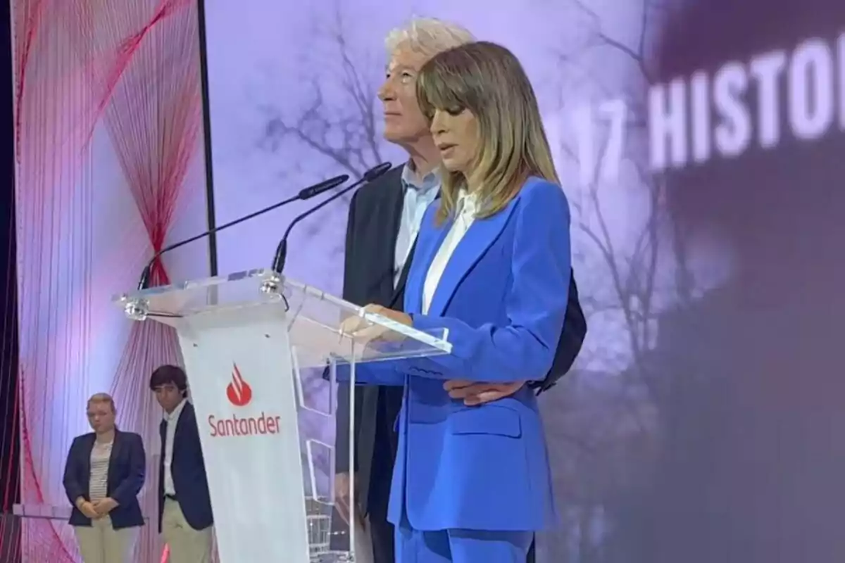 Dos personas de pie detrás de un atril con el logo de Santander, una de ellas leyendo mientras la otra la acompaña, en un escenario con fondo iluminado y dos personas más al fondo.