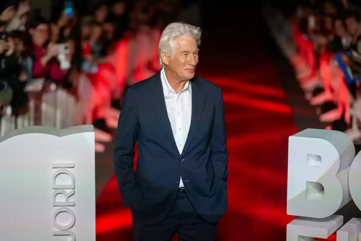 A gray-haired man in a dark suit walks down a red carpet surrounded by people taking his picture. A gray-haired man in a dark suit walks down a red carpet surrounded by people taking his picture.