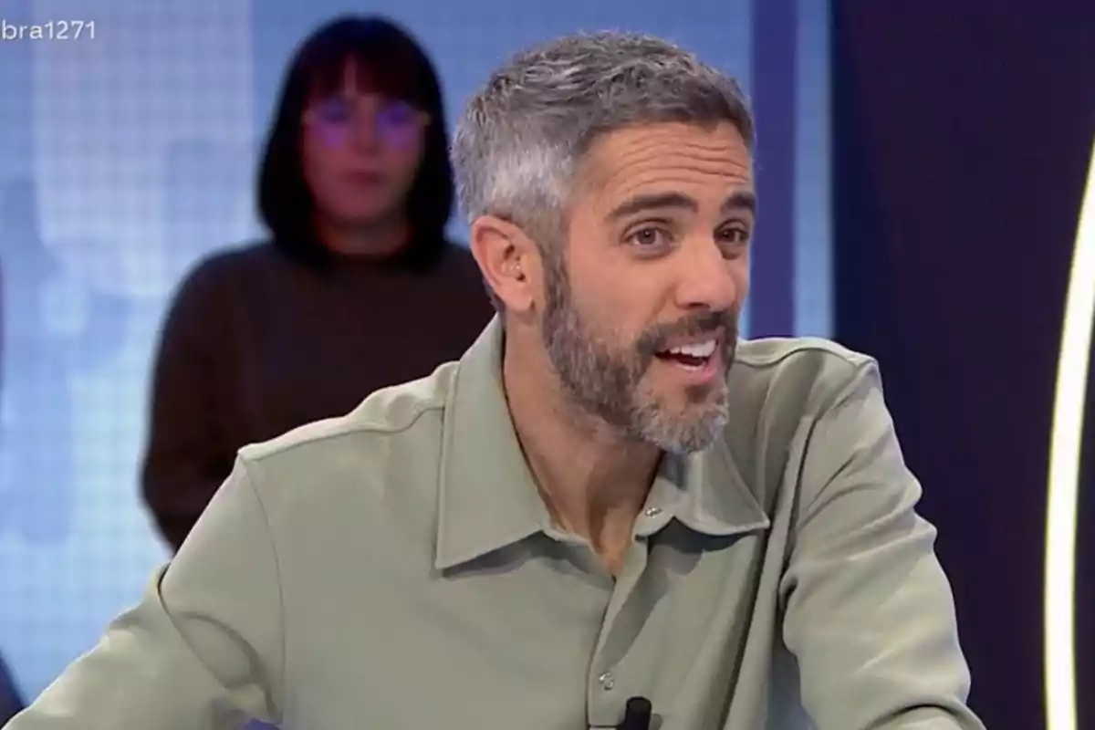 Un home amb barba i cabell canós somriu mentre està assegut en un estudi de televisió, amb una persona desenfocada al fons. Un home amb barba i cabell canós somriu mentre està assegut en un estudi de televisió, amb una persona desenfocada al fons.