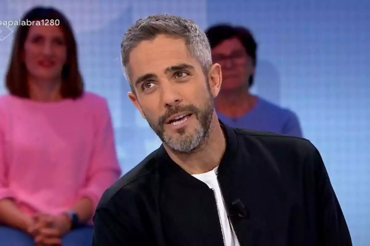Home de cabell curt i barba parlant en un programa de televisió amb dues persones assegudes al fons Home de cabell curt i barba parlant en un programa de televisió amb dues persones assegudes al fons