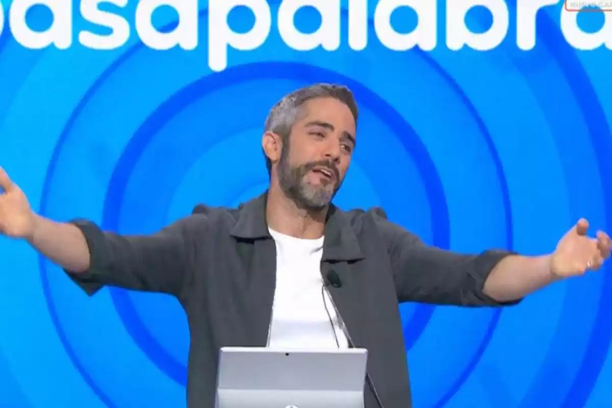 Home de cabells i barba canosos amb els braços estesos en un plató de televisió amb fons blau i lletres blanques Home de cabells i barba canosos amb els braços estesos en un plató de televisió amb fons blau i lletres blanques
