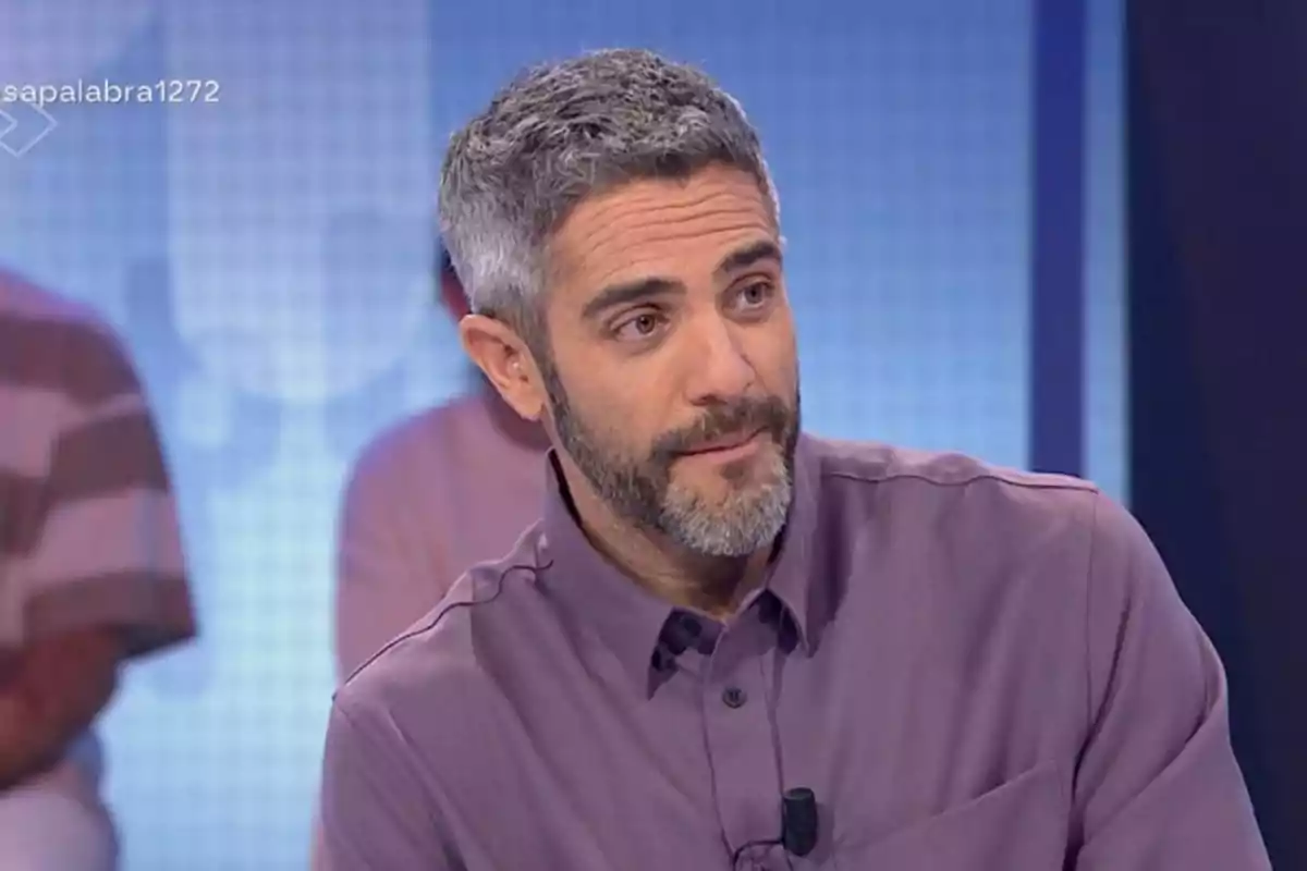 Un home amb barba i cabell canós, vestit amb una camisa morada, està assegut en un estudi de televisió. Un home amb barba i cabell canós, vestit amb una camisa morada, està assegut en un estudi de televisió.