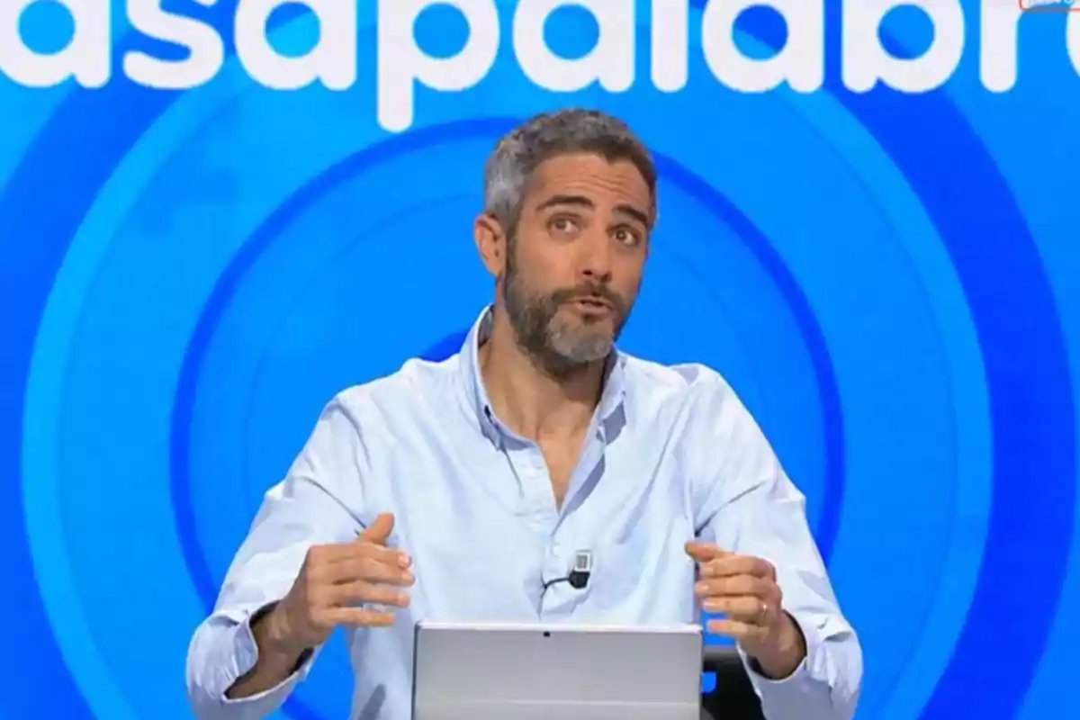 Home de cabell curt i barba parlant en un plató de televisió amb fons blau i lletres blanques Home de cabell curt i barba parlant en un plató de televisió amb fons blau i lletres blanques