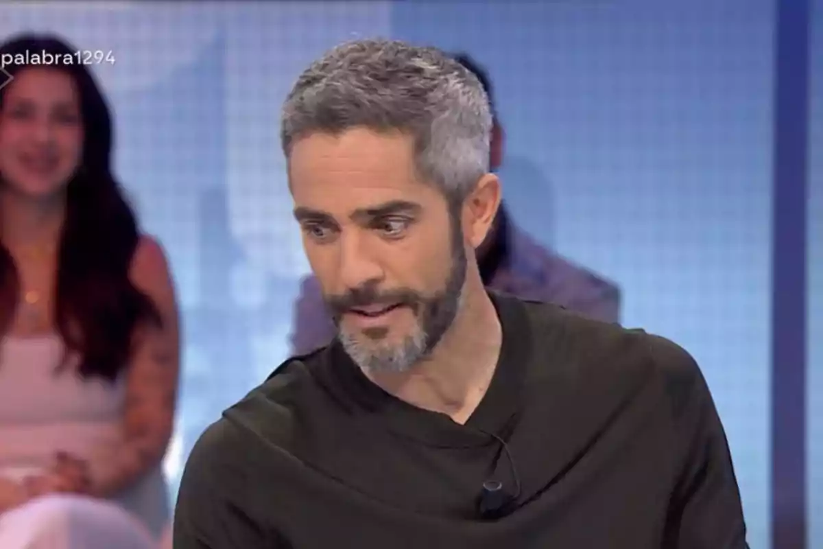 Home de cabells i barba canosos amb samarreta fosca participant en un programa de televisió amb persones desenfocades al fons Home de cabells i barba canosos amb samarreta fosca participant en un programa de televisió amb persones desenfocades al fons