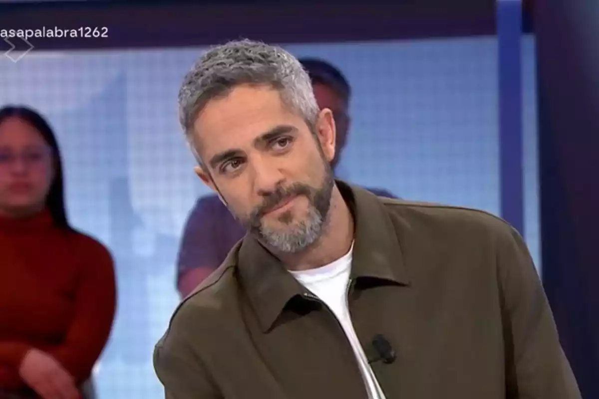 Un home amb barba i cabell canós és en un estudi de televisió, vestint una jaqueta verda i samarreta blanca, amb persones desenfocades al fons. Un home amb barba i cabell canós és en un estudi de televisió, vestint una jaqueta verda i samarreta blanca, amb persones desenfocades al fons.