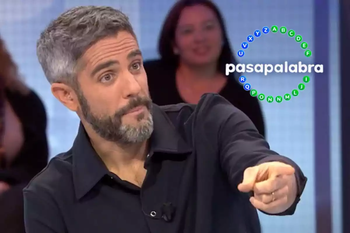 Un home amb barba i cabell canós assenyala cap endavant en un set de televisió amb el logotip de "Pasapalabra" al fons.