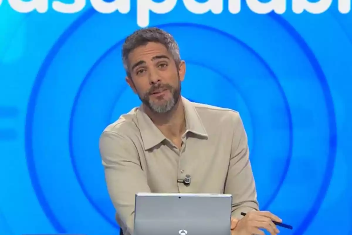 Un presentador de televisió amb barba i cabell curt està assegut davant d'un ordinador portàtil en un plató amb fons blau. Un presentador de televisió amb barba i cabell curt està assegut davant d'un ordinador portàtil en un plató amb fons blau.
