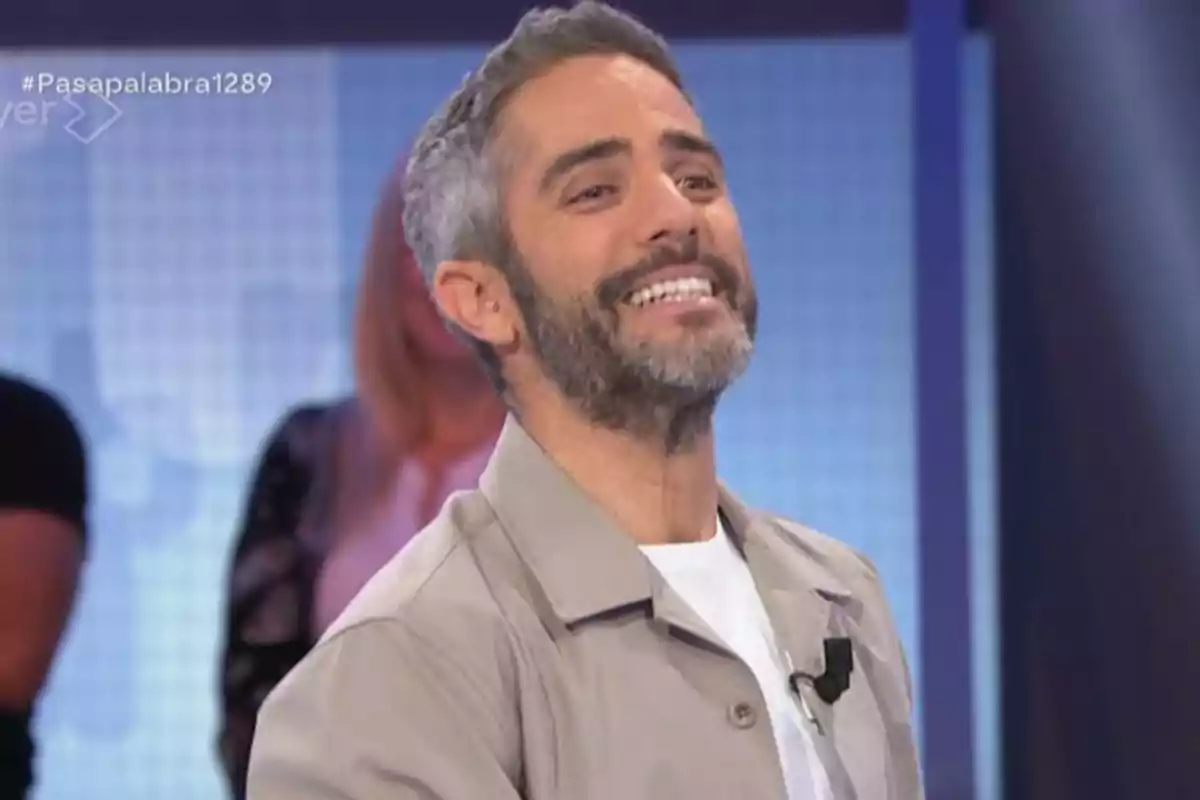 Home de cabell curt i barba somriu en un plató de televisió amb fons blau i persones desenfocades al darrere Home de cabell curt i barba somriu en un plató de televisió amb fons blau i persones desenfocades al darrere