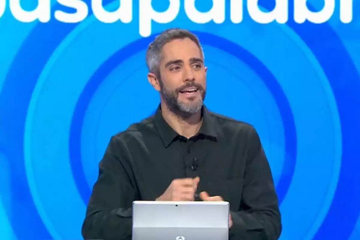 Un presentador de televisió amb barba i cabell curt està parlant davant d'una pantalla blava amb lletres blanques, mentre sosté un dispositiu electrònic.